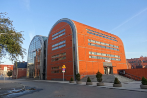 Ein großes oranges IBM-Hauptquartiergebäude mit zahlreichen Fenstern, umgeben von Straßeninfrastruktur, Fahrzeugen und Topfpflanzen unter einem bewölkten Himmel.