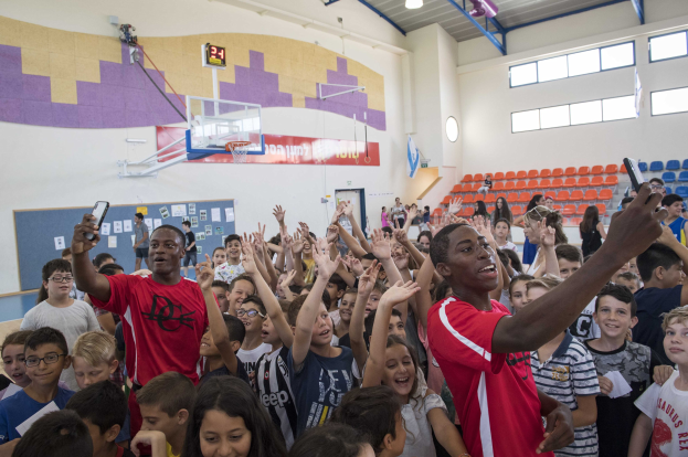 Kinder auf einem Basketballfeld mit Handys stehend, mit einer Anschlagtafel, Uhr, Torpfosten, Basketballnetz, Deckenleuchten, Stühlen und Fenstern im Hintergrund.