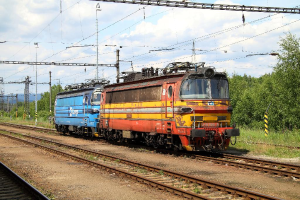 Eine Lokomotive auf Schienen mit Strommasten, Kabeln, Bäumen, Pflanzen und Wolken am Himmel.