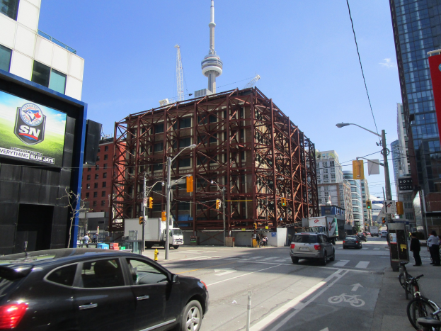 Eine belebte Stadtstraße mit Fahrzeugen, Fußgängern, parkenden Fahrrädern, Baustelle, Straßeninfrastruktur und dem CN Tower im Hintergrund bei klarem blauem Himmel.