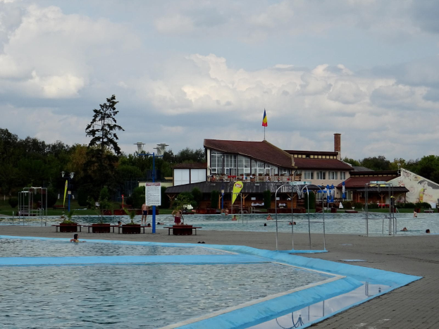 Ein großes Schwimmbecken mit Menschen darin, umgeben von Pfählen, Bänken, Topfpflanzen, einem Schild, einem Fahnenmast mit Flagge, einem Gebäude mit Fenstern, Straßenlaternen, einer Baumgruppe und einem bewölkten Himmel.