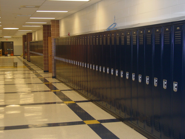 Eine lange Reihe von Metallspinden in einem Schulflur mit einem gefliesten Boden, einer Wand im Hintergrund, Deckenleuchten und ein paar verstreuten Gegenständen.