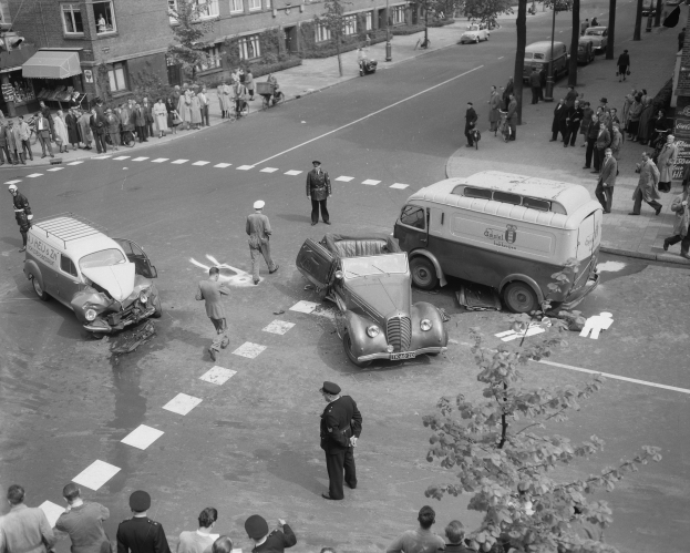 Schwarz-weiß-Szene eines Autounfalls an einer Straßenkreuzung mit mehreren Fahrzeugen, darunter ein Van, und Zuschauern; Gebäude, Bäume, Strommasten und Texttafeln im Hintergrund.