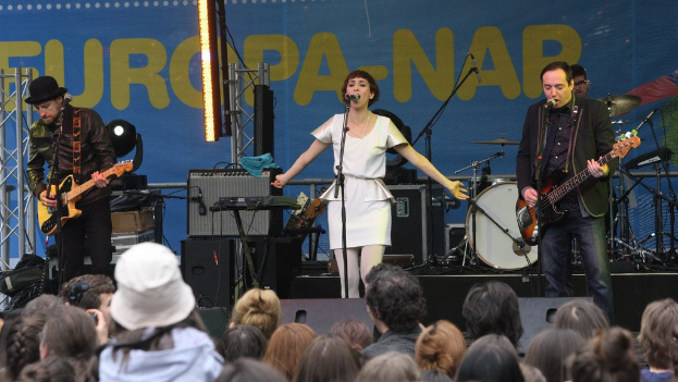 Vier Personen auf der Bühne: eine Frau in einem weißen Kleid, die singt und von zwei Männern mit Gitarren flankiert wird, vor einem Publikum und Lautsprechern an einer Wand im Hintergrund.