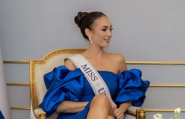 Frau in einem blauen Kleid und einem "Miss Universe 2019"-Band, die auf einem Stuhl sitzt und lächelt, umgeben von Blumen mit einer Wand im Hintergrund.