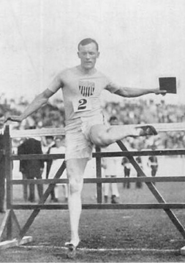 Schwarz-weißes Foto eines Mannes in Sportkleidung, der über ein Hindernis springt, mit Zuschauern im Hintergrund und dem Himmel darüber.