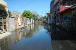 Ein enger Kanal in einer Stadt, gesäumt von Gebäuden auf beiden Seiten, mit ruhigem, klarem Wasser, das Bäume, Pfähle, Drähte und den Himmel spiegelt.