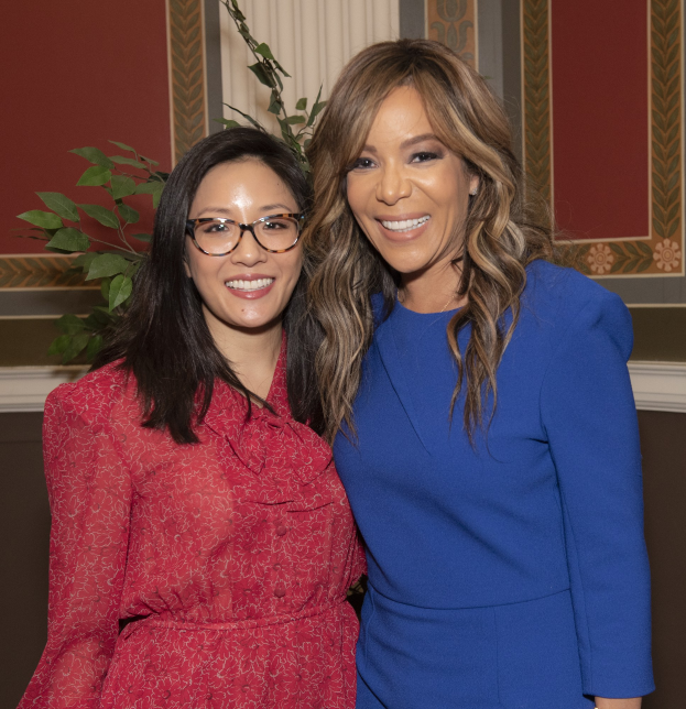 Zwei Frauen in Kleidern stehen lächelnd nebeneinander auf einer Veranstaltung, eine in Blau und eine in Rot mit Brille, mit einer Pflanze und einem Vorhang im Hintergrund.