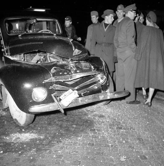 Schwarzes und weißes Bild eines schwer beschädigten Autos am Straßenrand mit eingedrückter Motorhaube und zersplitterten Fenstern, umgeben von einer Gruppe von Menschen, die die Szene begutachten.