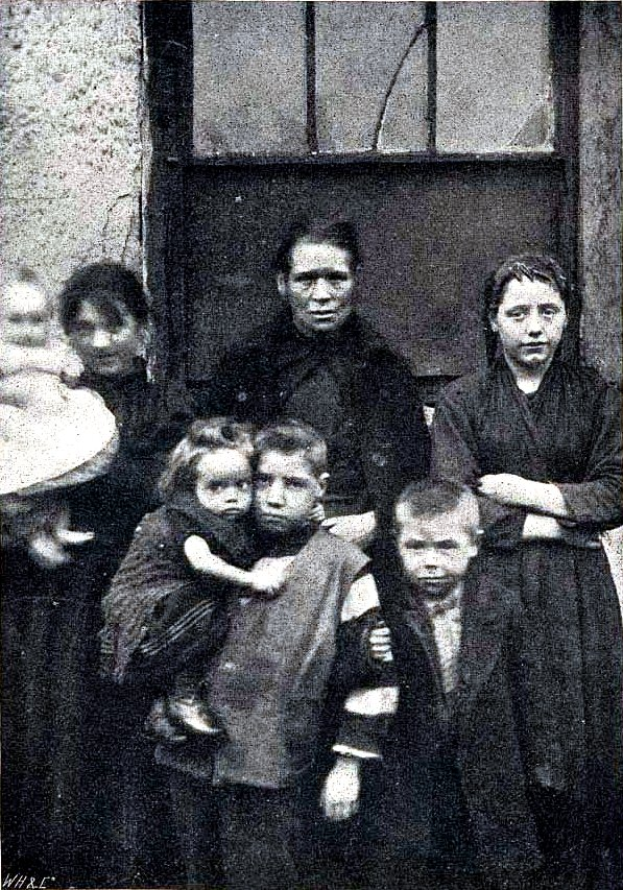 Ein altes Schwarz-Weiß-Foto einer Familie, die vor einer Wand mit einem Fenster posiert, mit Text unten, der "Mutter Teresa mit ihren Kindern" lautet.