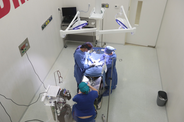 Eine Gruppe von Chirurgen führt einen Eingriff in einem Operationssaal durch, mit einem Patienten auf einer Trage umgeben von medizinischer Ausrüstung, einer textbedeckten Wand auf der linken Seite, einem Objekt auf dem Boden auf der rechten Seite und diverser medizinischer Ausrüstung im Hintergrund.