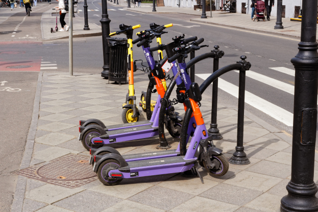 Eine Reihe von E-Scootern, die an einer Straße geparkt sind, mit Fußgängern und Radfahrern auf dem Gehweg, einem Müllcontainer im Vordergrund und Gebäuden, Bäumen und Pflanzen im Hintergrund.