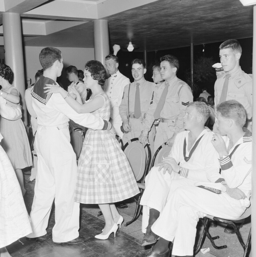 Schwarz-weißes Bild von Militärpersonal, das auf einer Tanzfläche tanzt und sich vergnügt, mit Säulen und Beleuchtung im Hintergrund.