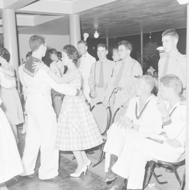Schwarz-weißes Bild von Militärpersonal, das auf einer Tanzfläche tanzt und sich vergnügt, mit Säulen und Beleuchtung im Hintergrund.