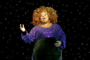 Eine Frau in einem lilafarbenen Kleid steht auf einer Bühne, beleuchtet von Hintergrundlichtern, als ob sie mitten in einer Vorstellung ist.