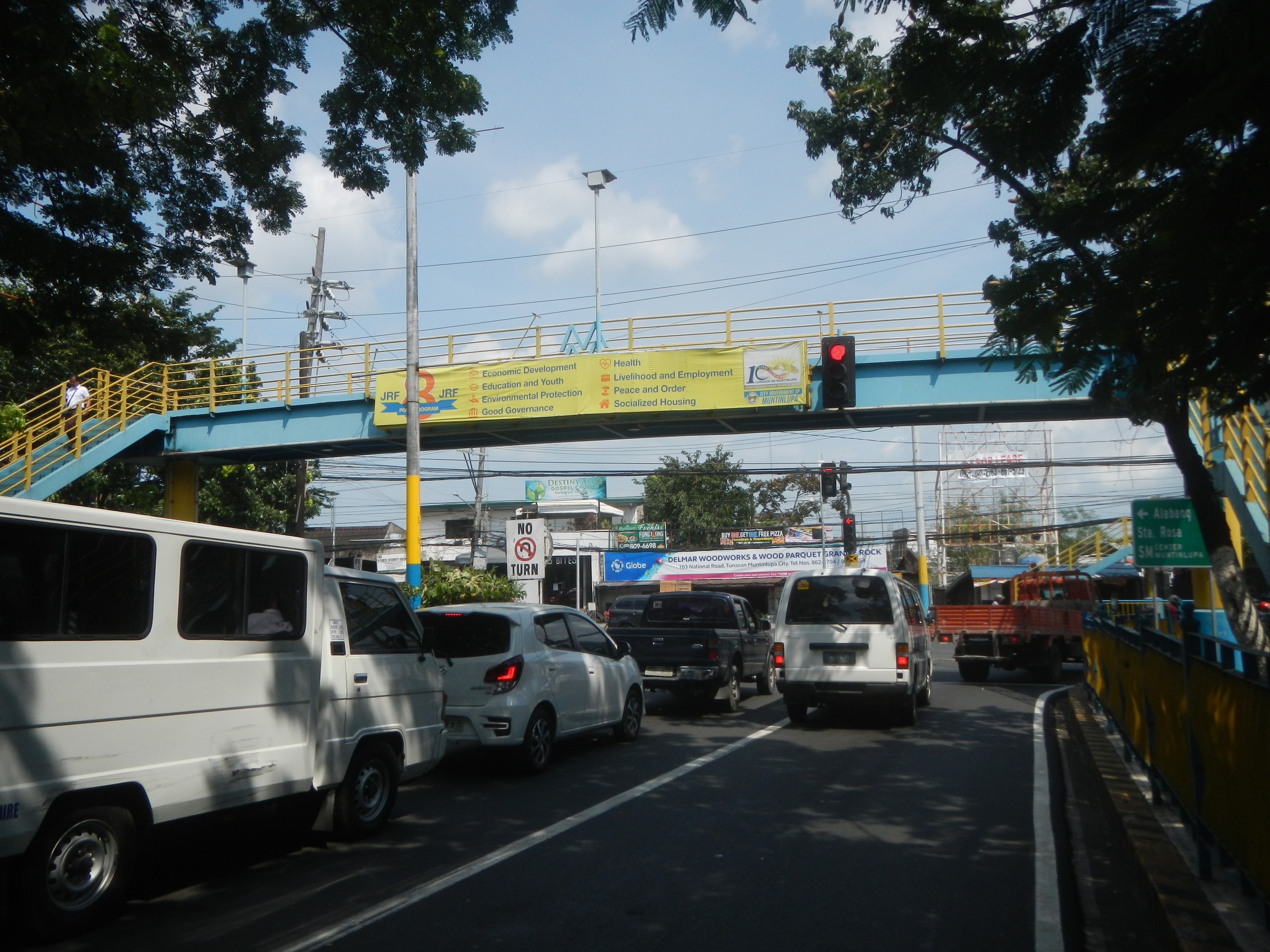 Vielbefahrene Straße mit Fahrzeugen, eine Brücke mit Geländern und Treppen, Laternen, Verkehrsampeln, Schilder, Bäume, Gebäude und ein bewölkter Himmel.