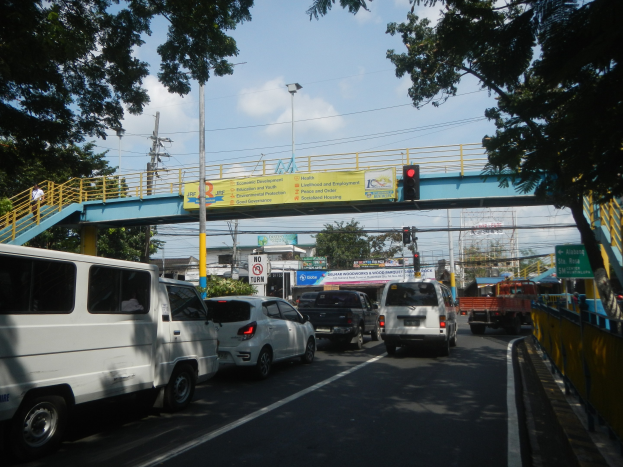 Vielbefahrene Straße mit Fahrzeugen, eine Brücke mit Geländern und Treppen, Laternen, Verkehrsampeln, Schilder, Bäume, Gebäude und ein bewölkter Himmel.