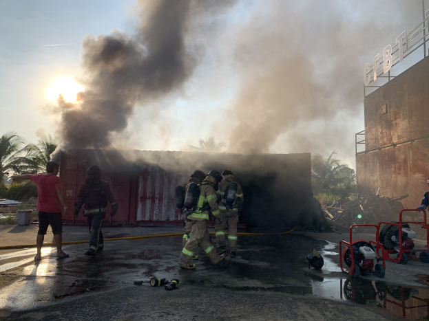 Feuerwehrleute mit Helmen arbeiten daran, ein Gebäudefeuer zu löschen, während dicker Rauch aufsteigt und in der Nähe ein weiteres Gebäude und Bäume zu sehen sind.