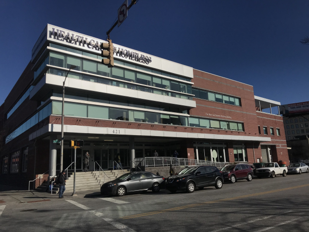 Großes Gebäude mit der Aufschrift "New York City Health Care Homeless Center" mit parkenden Autos, Fußgängern, Straßenmöbeln und Bäumen unter einem klaren blauen Himmel.