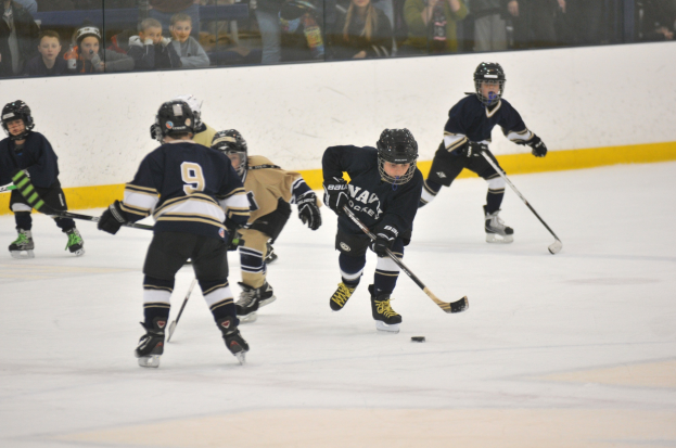 Gruppe junger Jungs in Helmen und Schlittschuhen, die Eishockey auf einer Indoor-Eisbahn spielen, jeder hält einen Hockeystock, mit sichtbaren Zuschauern durch eine Glaswand im Hintergrund.