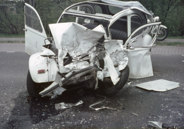 Ein Auto mit schwerer Frontschaden steht am Straßenrand, mit Bäumen, einem Gebäude und einem anderen Fahrzeug im Hintergrund.