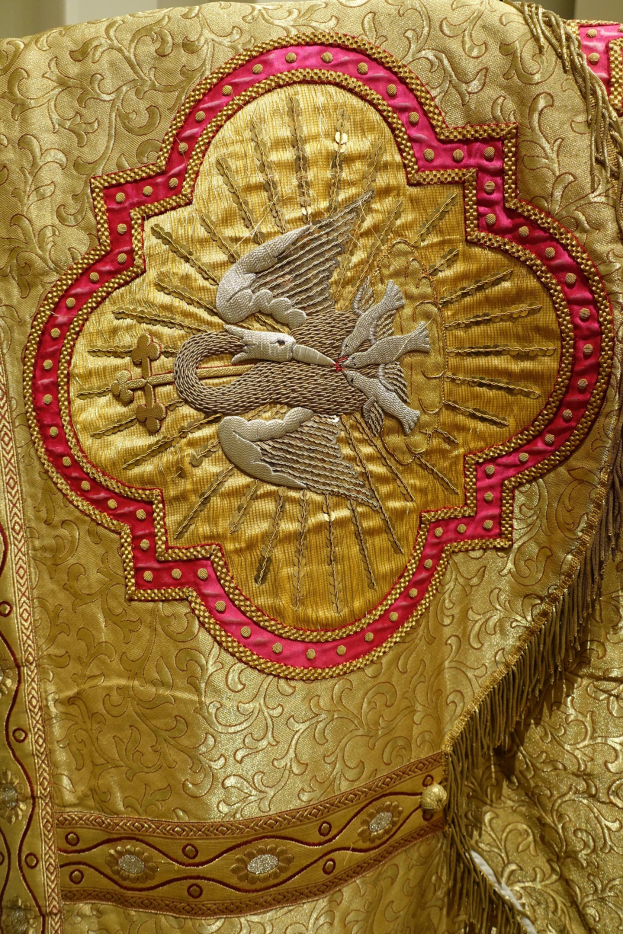 Nahaufnahme eines Priestergewandes mit aufwändiger Gold- und Rotstickerei und einem luxuriösen, königsähnlichen Design vor einer Wand im Hintergrund.