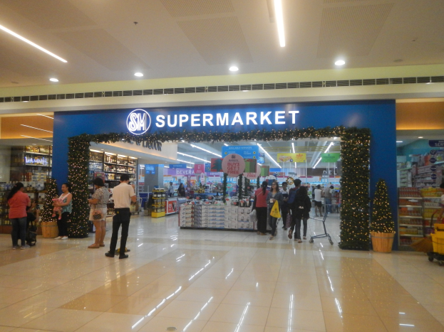 Eine Gruppe von Menschen, die vor einem Supermarkt stehen, einige halten Einkaufstaschen, mit einem Einkaufswagen, Topfpflanze, Regalen mit Gegenständen, Schildern und einer Decke mit Deckenleuchten.