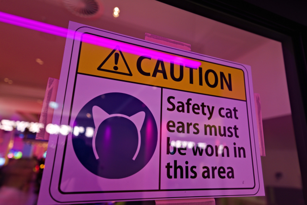 Hellgelbes Warnschild in einem Ladenschaufenster mit schwarzem Text "Warnung: Sicherheits-Katzenohren müssen in diesem Bereich getragen werden."