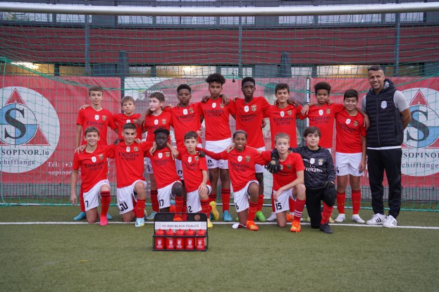 Gruppe junger Männer in roten T-Shirts und schwarzen Shorts auf einem Fußballfeld mit einer Kiste roter Gegenstände davor, umgeben von Bannern mit der Aufschrift "FC Rot-Schwarz Eglaute - Champions League" und Stadion-Elementen im Hintergrund.