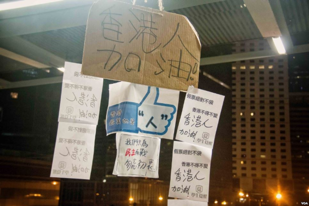 Menschen, die vor einem beleuchteten Gebäude in der Nacht mit Schildern protestieren, die von der Decke hängen.