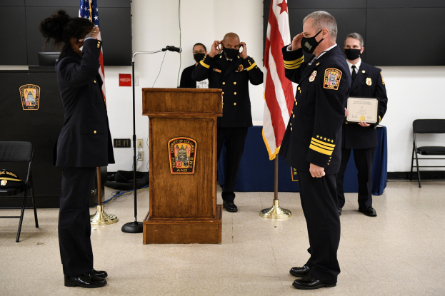 Feuerwehrleute in Helmen stehen auf einem Dach und halten Schlauchleitungen, mit Geländern und Treppen darunter und einem Gebäude und Himmel im Hintergrund.