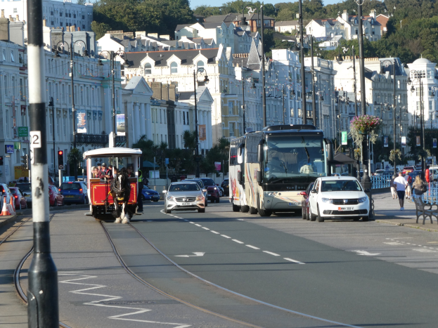 Eine belebte Stadtstraße mit hohen Gebäuden, Fahrzeugen, Fußgängern, Verkehrszeichen, Straßenlaternen, Schildern, Bäumen und einem klaren blauen Himmel.