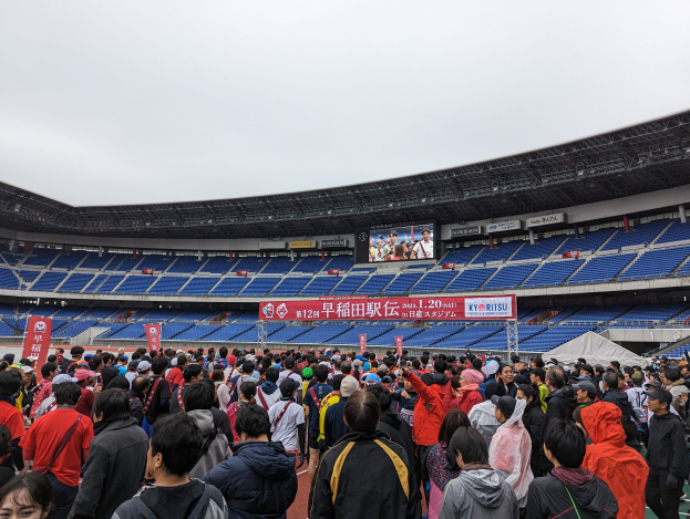 Große Menschenmenge vor einem Stadion während der Eröffnungsfeier der Olympischen Spiele Tokyo 2020, mit Bannern, Stühlen, einem Bildschirm und einem sichtbaren Himmel im Hintergrund.