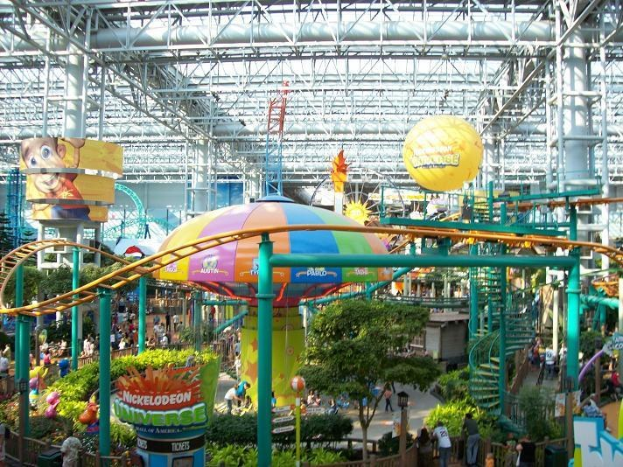 Ein großer Indoor-Wasserpark mit einer Achterbahn, umgeben von Menschen, Bäumen, Pflanzen und einer einladenden Beleuchtung.