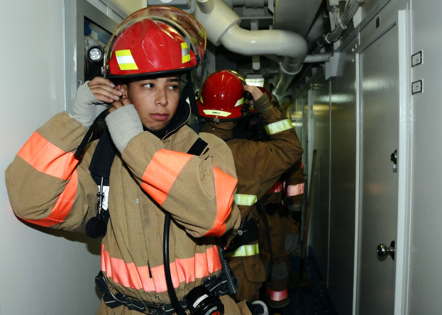 Feuerwehrleute in Uniform in einem Übungsraum mit Rohren und Ausrüstung im Hintergrund.