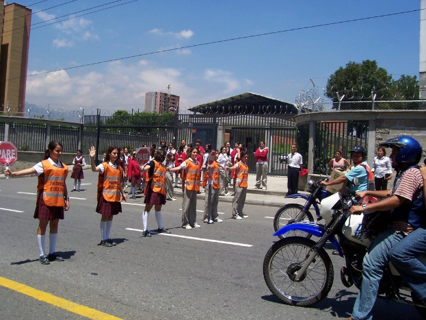Eine Gruppe von Schülerinnen in Uniform hält Fußgänger auf einer Straße an, mit Fahrradfahrern rechts, die Helme tragen, eine Schülerin links mit einem Schild und einem Hintergrund aus Zaun, Gebäuden, Bäumen und Himmel.