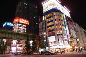 Eine befahrene Straße mit fahrenden Autos, ein Gehweg mit Fußgängern, Bäume und Ampelmasten, Gebäude mit Texttafeln, eine Werbetafel und Lichter oben, unter einem dunklen Himmel.