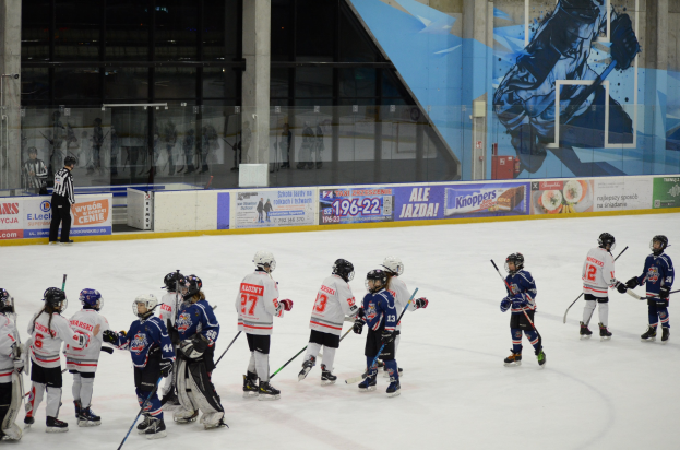 Gruppe junger Menschen beim Eisschlittschuhlaufen auf einer Indoor-Eisfläche, die Helme tragen und Stöcke halten, mit einer Person auf der linken Seite und Bannern, Säulen und Glaswänden im Hintergrund.