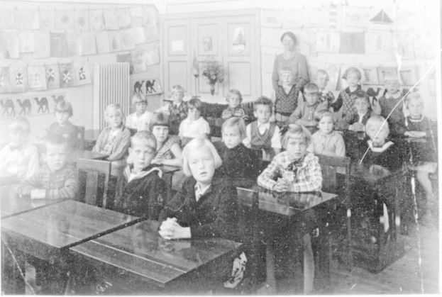 Schwarzes und weißes Foto einer Klasse mit Kindern auf Bänken sitzend und stehend, mit einem Blumentopf, Papieren an einer Tafel, einer Tür und Deckenlampen an der Rückwand.