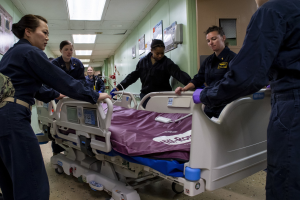 Medizinisches Personal in Uniformen um ein Krankenhausbett in einem Raum mit Wandplakaten und Deckenleuchten versammelt.