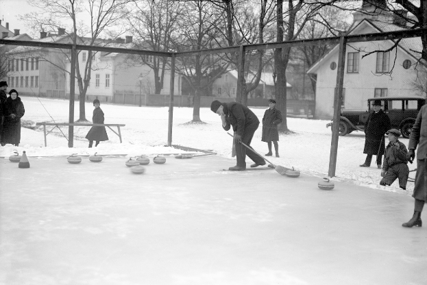 Schwarzes Bild von Menschen, die Curling auf einer Eisfläche spielen, umgeben von einem Zaun, einer Bank, Bäumen, Gebäuden und einem geparkten Fahrzeug im Hintergrund.