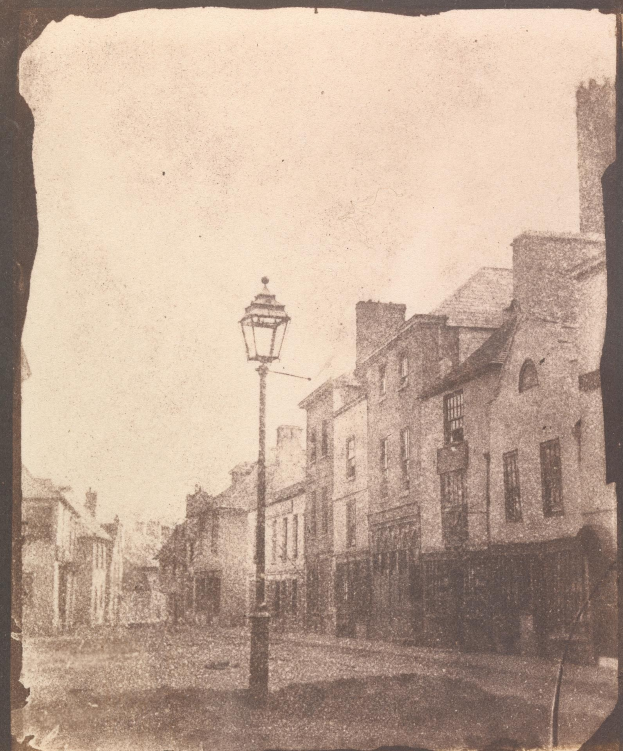 Alte Schwarz-Weiß-Fotografie einer Straßenlaterne an einer Kreuzung, flankiert von mehrstöckigen Gebäuden unter einem wolkenlosen Himmel.