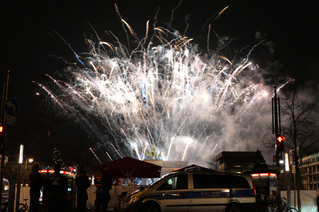Polizeiauto vor einer Menschenmenge während einer Silvesterfeier in Berlin geparkt, mit farbigen Feuerwerken, die den Himmel und Gebäude erleuchten.