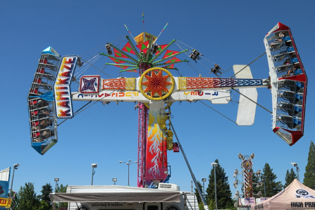 Ein Karussell auf einem Volksfest mit Menschen darauf, umgeben von Zelten, Bannern, Pfählen, Lichtern, Bäumen und anderen Gegenständen, mit dem Himmel im Hintergrund.