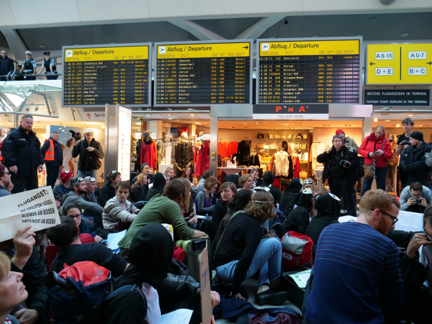 Eine große Gruppe von Menschen sitzt und steht in einem Flughafen während einer Protestaktion, mit Gepäck und Informationsschildern im Hintergrund.