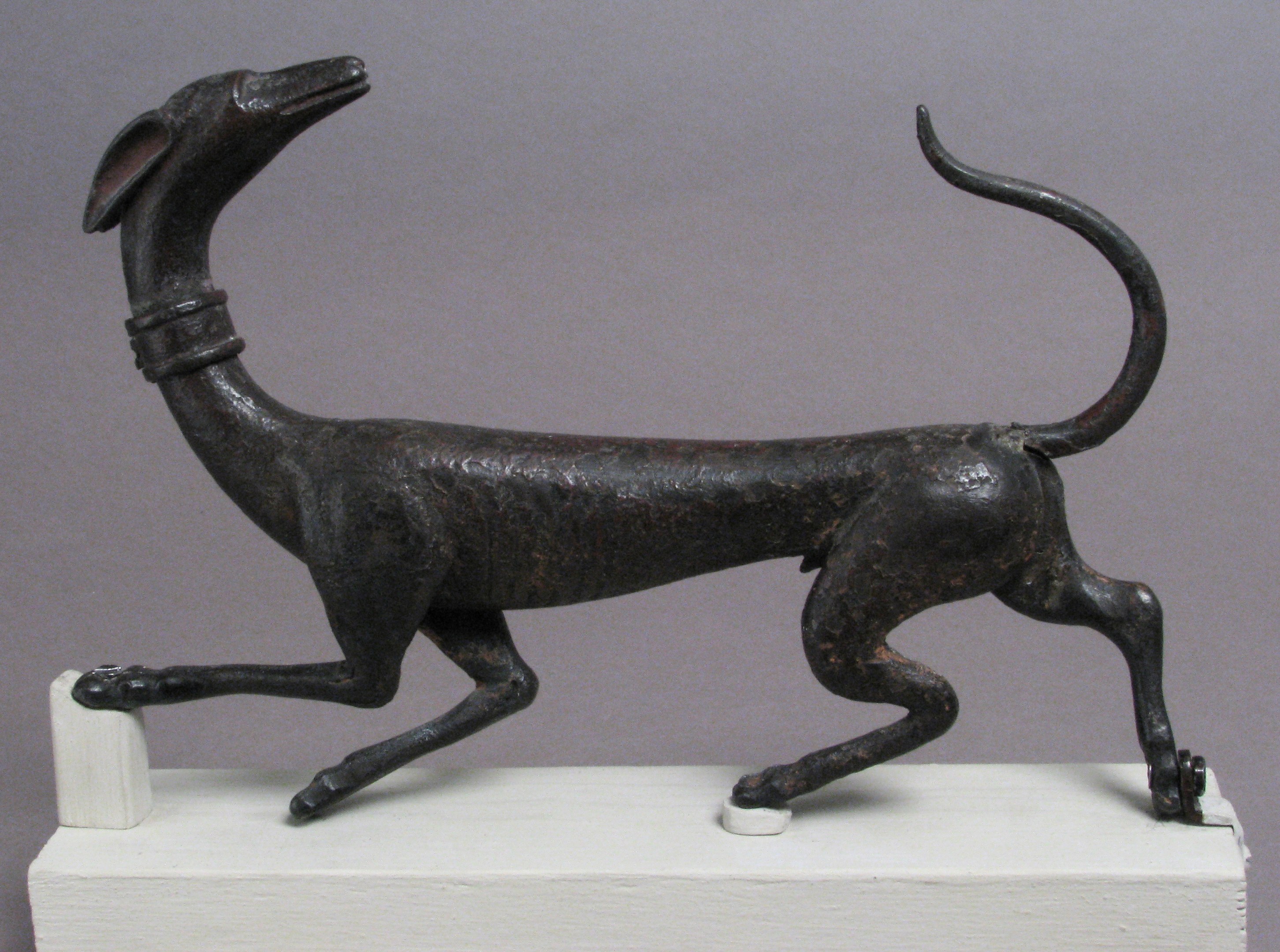 Eine Bronzestatuette eines Greyhounds auf einem weißen Sockel vor einem grauen Hintergrund mit detailliertem Fell und Zügen.