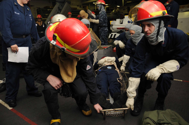 Rettungsteam-Mitglieder auf dem Deck eines Schiffs helfen bei einer Trage, einer hält Dokumente, während ein anderer die Ausrüstung verwaltet, mit einer Tasche und Hintergrundgegenständen sichtbar unter Deckenlampen.