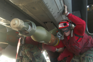 Eine Gruppe von Männern in roten Jacken und Tarnuniformen arbeitet an einem Flugzeug, wobei einer der Männer einen Helm, eine Schutzbrille und eine Uhr trägt und im Hintergrund eine Rakete zu sehen ist.