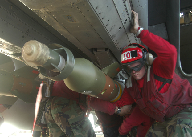 Eine Gruppe von Männern in roten Jacken und Tarnuniformen arbeitet an einem Flugzeug, wobei einer der Männer einen Helm, eine Schutzbrille und eine Uhr trägt und im Hintergrund eine Rakete zu sehen ist.