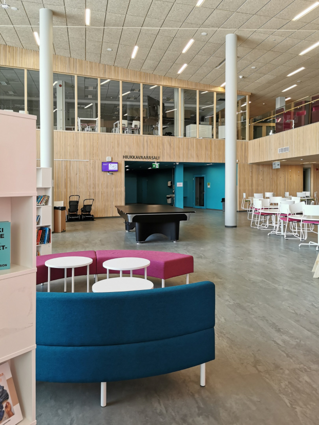 Großer Raum, der an eine Schulbibliothek erinnert, mit mehreren Tischen, Stühlen, Sofas, Hockern, Bücherregalen, Deckenleuchten, Säulen und einem wandmontierten Fernseher.